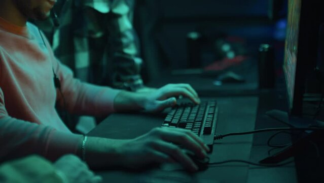 Medium shot of young Arab guy sitting at computer in cyber cafe, putting on headphones and preparing to play fun cartoon video game, while diverse friends are standing around and watching