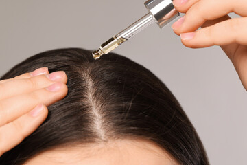 Woman applying essential oil onto hair roots on light grey background, closeup