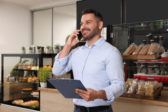 Business owner with clipboard talking on phone in his cafe - Powered by Adobe