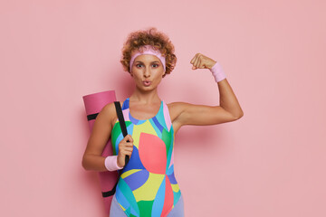 Close up shot of young woman dressed in gymnastic sportswear with yoga mat poses on pink background with one fist up, barbie fashion concept, copy space