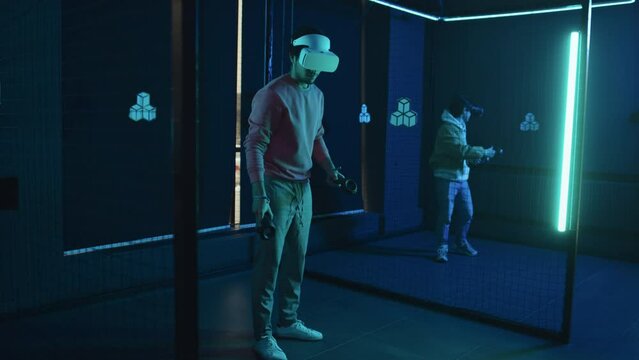 Full shot of two young men in VR headsets playing augmented reality fighting games in empty dark arena with blue neon light, slashing, hitting and striking simulated three-dimensional objects