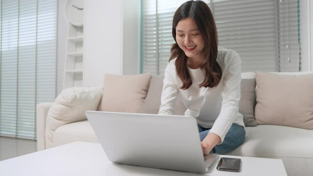 Asian teenage girl typing laptop computer working sitting on sofa in living room at home. Cheerful asian woman work freelance using computer notebook answer chat from customer at home office.