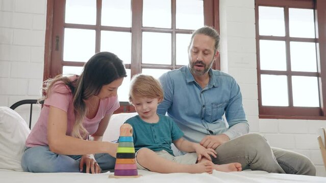 Happy caucasian parents playing wooden toys with little son together in bedroom at home. Father and mother play and teasing with child on the bed in holiday. Family relationship concept.