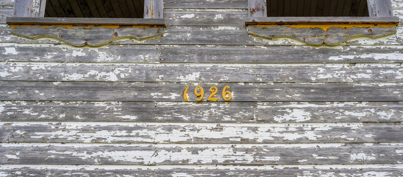 Image of an old, dilapidated building with pealing white paint and the date 1926 in yellowish-orange numbering. The bottom of two windows can be seen at the top of the photo.
