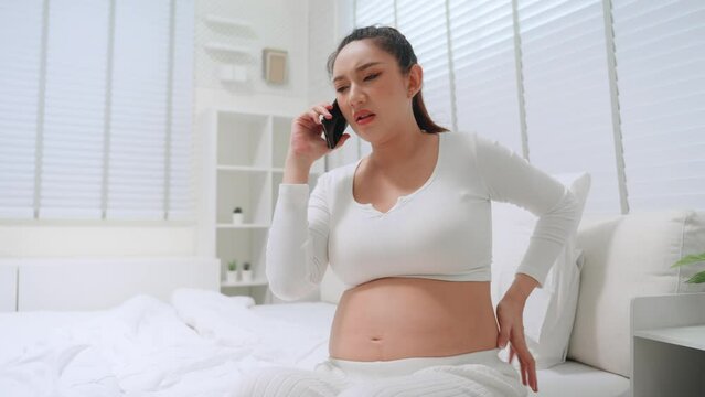 Pregnant asian woman calling with mobile phone and ask to advice the doctor have symptom backache effect from pregnancy at home. Maternity prenatal care and pregnancy concept.