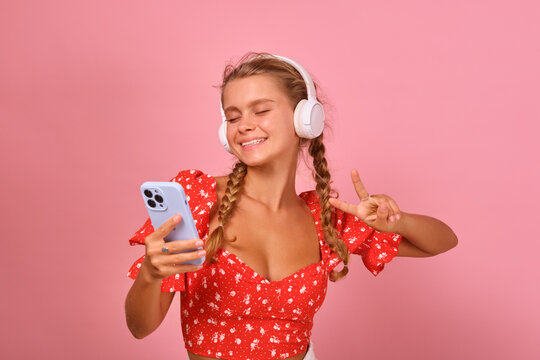 Young energetic positive Caucasian woman dances enjoying listening to music in wireless headphones and holding mobile phone with application for watching video clips stands in pink studio.