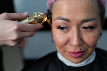 The hairdresser shaves the temple of a female client. Asian woman with short pink hair in barbershop.