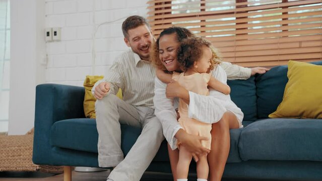 Happy parents embracing and teasing little daughter in living room at home. Father and mother using spare time with baby together at warmth house. Family relationship concept.