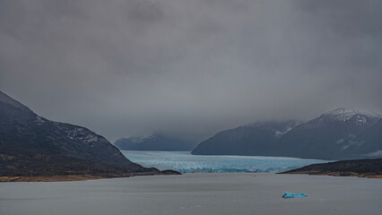 The blue glacier of Perito Moreno stretches between the mountain slopes. Breakaway icebergs float in the lake. Cloudy and foggy. Argentina. El Calafate. Lago Argentino.