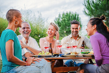 Mature people enjoying at lunch party drinking red wine sitting at dining table at their home backyard. Social gathering of middle aged best friends, having fun and talking on a funny birthday reunion