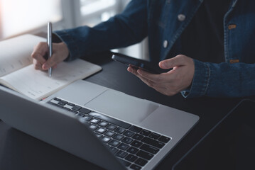 Business woman sitting at table using mobile phone, online working on laptop and making notes in...