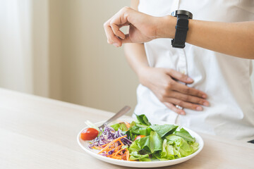 Intermittent fasting, health care asian young woman dietary, having stomach ache, temptation hungry of breakfast food in morning on table at home, looking at watch on her wrist but not yet time to eat