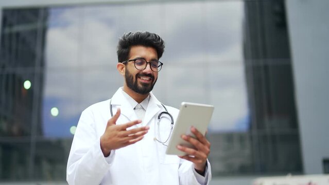 Happy young doctor talking online via video call, holding a tablet and standing outside in front of a hospital building. Medical worker physician in a white coat and glasses consults a patient