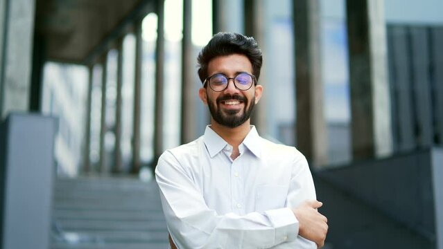 Portrait of a young bearded businessman smiling and looking at the camera. Handsome male in a white shirt and glasses posing with crossed arms on the street in front of an office building. Head shot