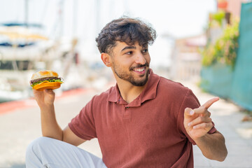 Young Arabian handsome man holding a burger at outdoors pointing to the side to present a product