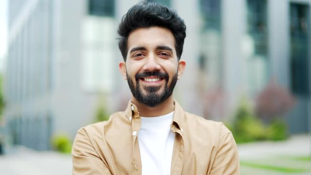 Close up. Portrait of a young bearded man smiling and looking at the camera. Handsome positive male posing standing on the street near the office building. Head shot of optimistic friendly businessman