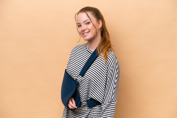 Young caucasian girl with broken arm and wearing a sling isolated on beige background smiling a lot