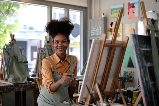 Portrait of smiling young woman with Afro haired wearing apron painting picture with brush on easel in art studio
