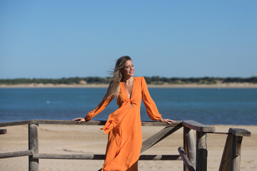 Pretty young blonde woman in orange dress leaning on the wooden railing of the beach promenade in the wind. In the background on the horizon blue sky and sea. Holiday and travel concept. © @skuder_photographer