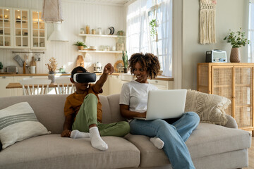 African American family mom and son sit on sofa with electronic devices. Mother-parent sit with...