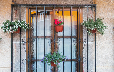 Ventana con cristal y rejas donde cuelgan unas macetas con plantas en flor. Casa de arquitectura moderna, pero tradicional en Hoyos del Espino, Ávila, España. 