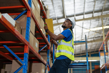 Black man warehouse worker wearing vest and helmet safety look at clipboard and checking product number on box in shelf at warehouse factory store. Logistics, Distribution Center concept
