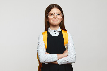 Proud nerdy pupil girl wearing eyeglasses smiling and looking at camera with hands crossed while...
