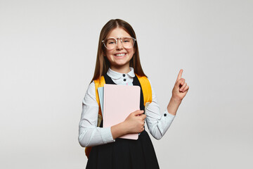 Clever little nerdy girl kid in school uniform and eyeglasses holding books and pointing up,...