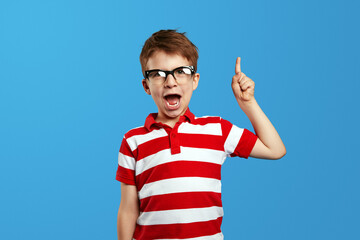 Fototapeta premium Little smart nerdy boy in eyeglasses and red striped shirt shouting and pointing up against blue background. Studio portrait of cute preschool child