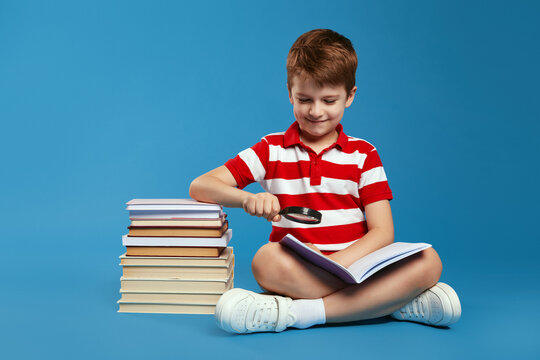 Happy child in casual clothes smiling and using magnifying glass to read book while sitting crossed legged on floor near bunch of books, isolated over blue background