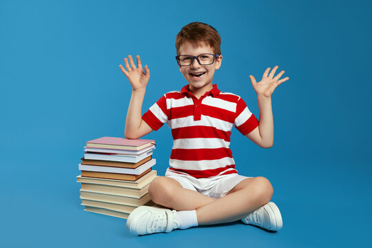 Smart schoolboy in red striped shirt and eyeglasses, celebrating deadline, finished reading all the books for the summer, sitting crossed legged on floor near bunch of books, isolated over blue wall