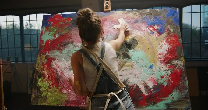 Millennial girl artist paints picture in loft studio, applying paint to canvas with spatula. Abstract cinematic. Fully concentrated sophisticated female artist is drawing picture on easel. Slow motion
