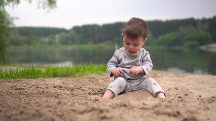 Naklejka na ściany i meble A little boy sits on the beach by the river and plays in the sand. A two-year-old child plays in the sand against the background of the river.