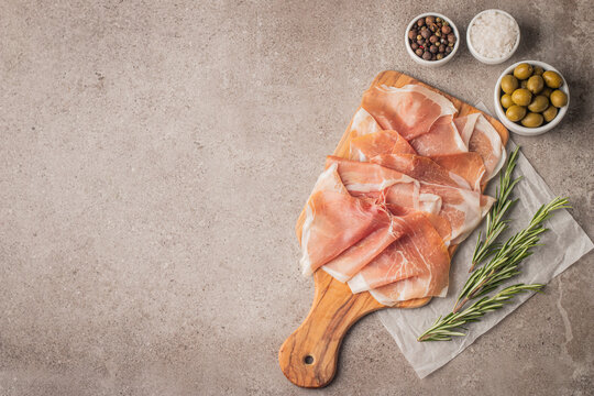 Thin slices of Italian prosciutto with green olives, spices and rosemary on wooden cutting broad. Raw dried meat.