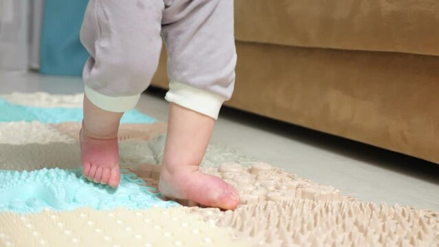 Barefoot toddler girl in grey pants walking around house for first time. Small legs of daughter step on colorful soft carpet near couch, close-up