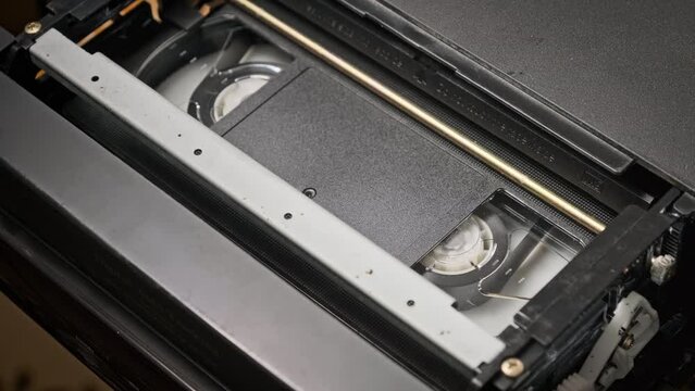 VHS cassette is played inside a VCR tape recorder, top view. Reels of videotape rotate. Video cassette with a blank tag is starting playback. Old video recorder inside close-up. Playing old movie.