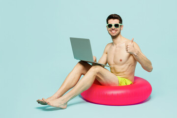 Full body young IT man wear green shorts swimsuit relax near hotel pool sit on rubber ring work on laptop pc computer show thumb up isolated on plain blue background. Summer sea rest sun tan concept.