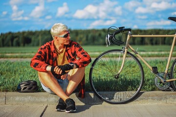 a blond guy in glasses and a red shirt, taking off his bicycle helmet, sits on the side of the road near a vintage bike