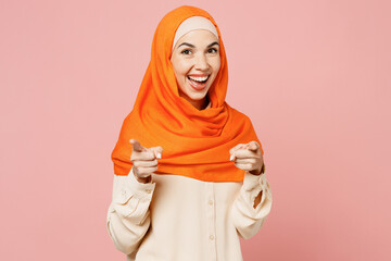 Young fun arabian asian muslim woman wear orange abaya hijab point index finger camera on you motivating encourage isolated on plain pink background studio. Uae middle eastern islam religious concept.