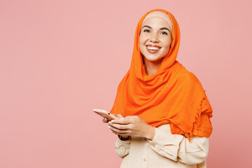 SIde view young arabian asian muslim woman wear orange abaya hijab hold in hand use mobile cell phone isolated on plain light pink background studio. People uae middle eastern islam religious concept.