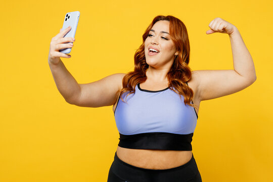 Young chubby overweight plus size big fat fit woman wear blue top warm up train do selfie shot mobile cell phone show muscles isolated on plain yellow background studio home gym Workout sport concept