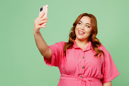 Young chubby overweight redhead woman in casual clothes pink dress doing selfie shot on mobile cell phone post photo on social network isolated on plain light green color background Lifesyle concept