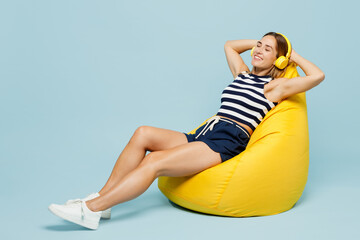 Obraz premium Full body young woman she wear striped tank shirt casual clothes sit in bag chair listen to muisc in headphones close eyes isolated on plain pastel light blue cyan background studio Lifestyle concept