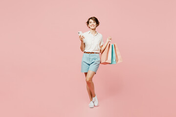 Full body young happy woman wear casual clothes hold shopping paper package bags hold in hand use mobile cell phone isolated on plain pink color background studio. Black Friday sale buy day concept.