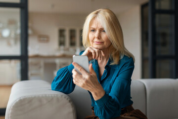 Upset european mature woman sitting on couch at home, holding cell phone, looking at gadget screen,...