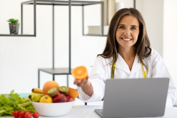 Smiling mature caucasian lady doctor nutritionist in white coat at table with laptop with organic fruits show orange