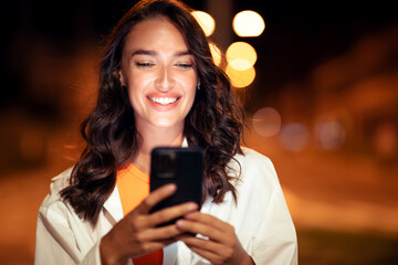 Cityscape and smiles. Portrait of happy young lady walking in evening city and using cellphone, free space