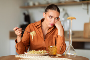 Upset caucasian woman eating tasteless pasta or needs to keep diet, sitting at table in kitchen interior