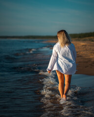 A beautiful woman in a white shirt with slender legs moves along the beach along the ocean