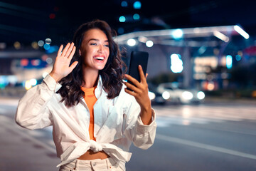 Excited european woman using cellphone having video call while walking in the city at night, waving hello at webcamera